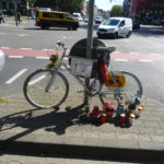 Ein nicht mehr fahrtüchtiges Fahrrad, weiss angestrichen angekettet am Hansemannplatz in Aachen. Vor dem Fahrrad stehen Kerzen. Im Hintergrund, Autos