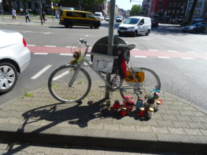 Ein nicht mehr fahrtüchtiges Fahrrad, weiss angestrichen angekettet am Hansemannplatz in Aachen. Vor dem Fahrrad stehen Kerzen. Im Hintergrund, Autos