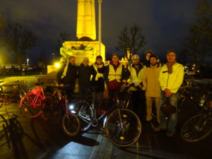 Posiertes Foto: 8 Personen in einer Reihe, vor ihnen mehrere Fahrräder, im Hintergrund der untere Teil der gëlle Fra. Alle winterlich gekleidet, die meisten in heller Kleidung, einige mit Fahrradhelm