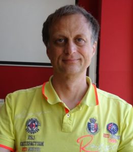 Foto des Autors von 2015. Gelbes Polohemd mit einigen Badges drauf, Hintergrund rot mit weisser Ecke.