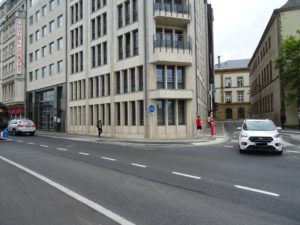 Ein Eckgebäude, davor Fussweg mit 2 Fussgängern und einem Hochbordradweg mit Schild. Ein Abbiegendes Auto von vorne