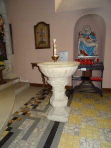 Der Taufstein, im Hintergrund in einer Nische die Siebenschmerzenmaria, davor ein Ständer mit Kerzen, links davon eine Osterkerze