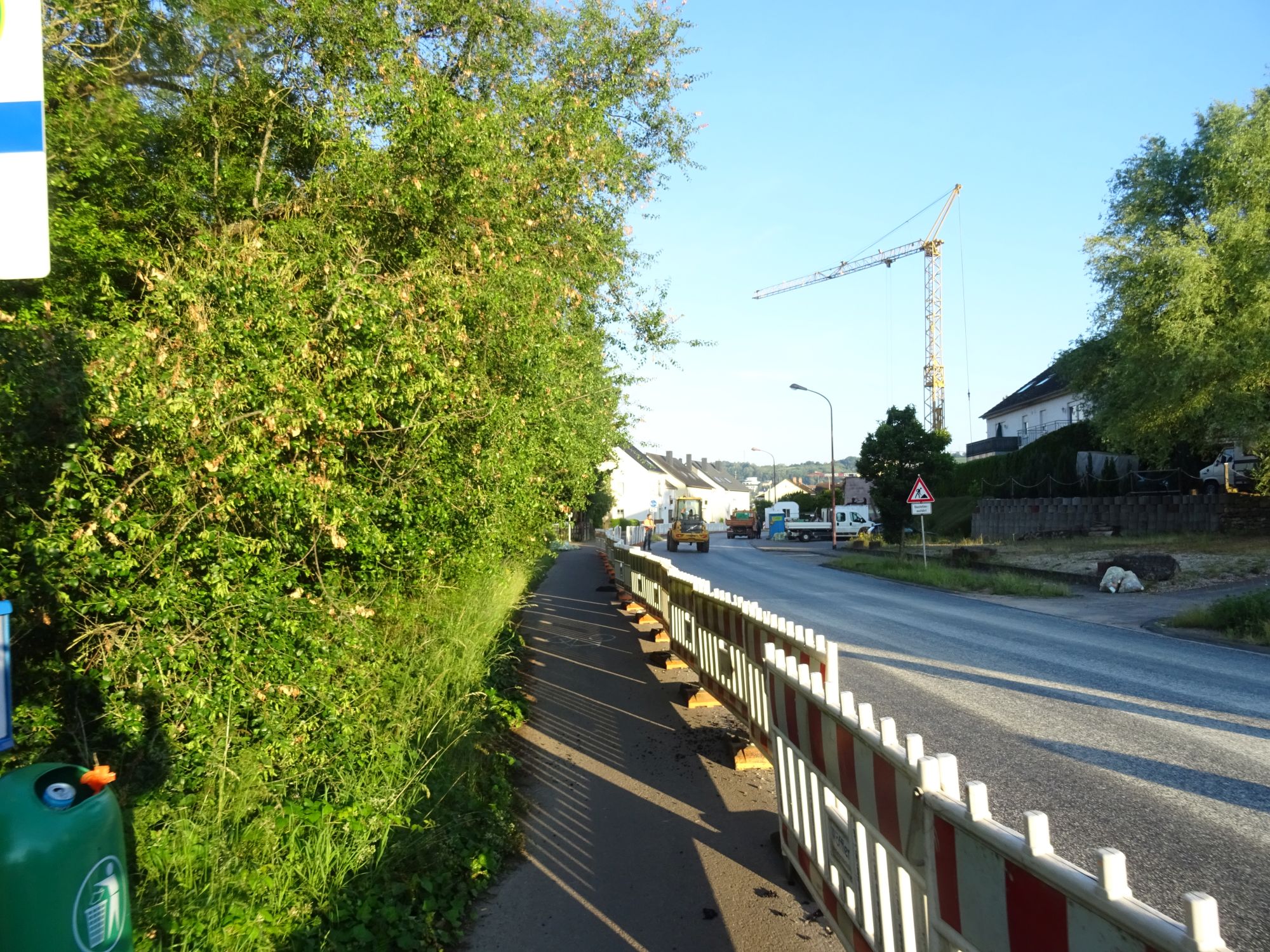 Links Bäume, Mitte Radweg mit Bauzaun, rechts die noch nicht freigegebene Fahrbahn, im Hintergrund ein Baufahrzeug